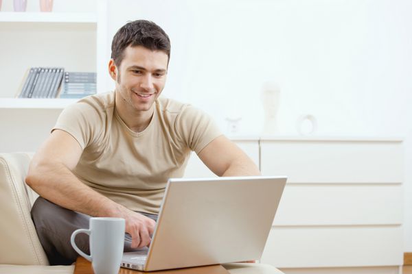 مرد جوان شادی با تی شرت نشسته روی مبل خانه روی لپ تاپ کار می کند و لبخند می زند