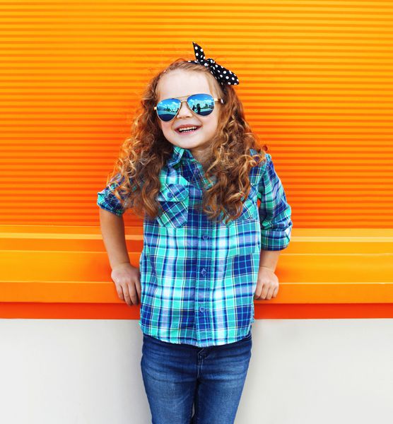 مفهوم بچه مد - کودک دختر کوچک شیک با پیراهن و عینک آفتابی مقابل دیوار نارنجی رنگارنگ