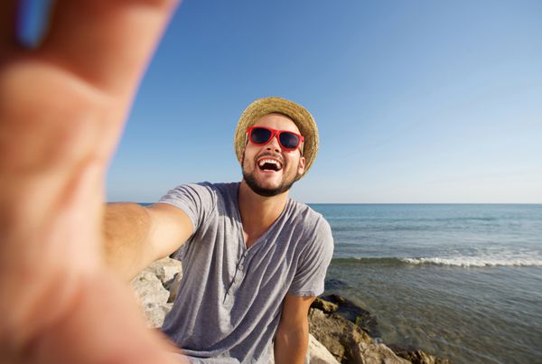 مرد خوشحال در تعطیلات در حال خندیدن در ساحل در حال گرفتن سلفی