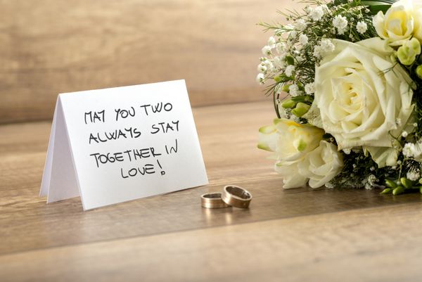 کارت تبریک عروسی از نزدیک همراه با پیام عاشقانه دسته گلهای سفید تازه و یک جفت حلقه در بالای میز چوبی