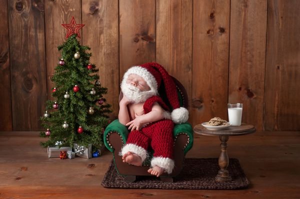 دو هفته ای نوزاد پسری که کت و شلوار بابانوئل قلاب بافی با ریش به تن دارد او روی صندلی راحتی می خوابد در استودیو با وسایلی مانند درخت کریسمس لیوان شیر و کلوچه های قلاب بافی عکس گرفته شده است