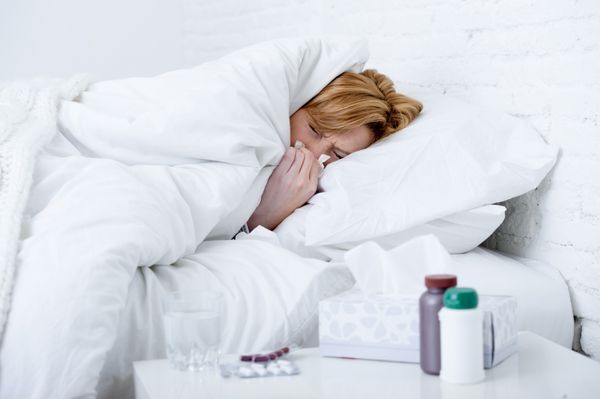 زن بیمار جوان با بینی عطسه دمیدن در بافت دراز کشیده روی تخت مبتلا به سرماخوردگی زمستانی و علائم ویروس آنفولانزا با داشتن قرص و قرص دارو در بیماری و مفهوم مراقبت های بهداشتی خانگی