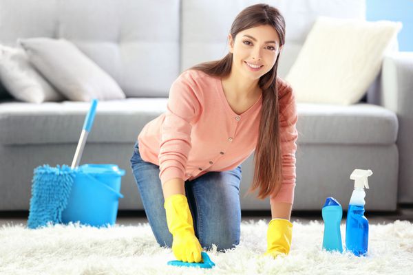 زن زیبا با دستکش محافظ فرش را با برس و اسپری تمیز می کند