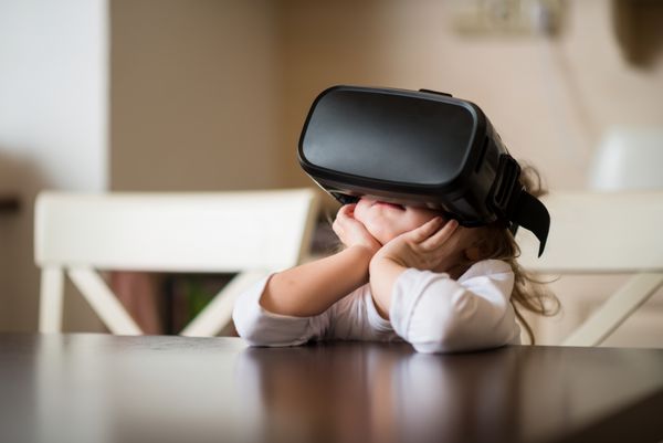کودکی با هدست واقعیت مجازی که پشت میز داخل خانه نشسته است