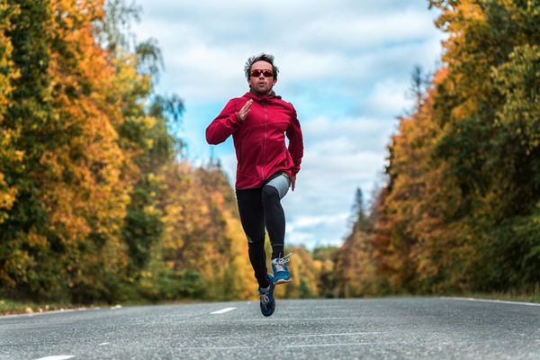 مردی با لباس ورزشی و عینکی که در جنگل پاییزی در جاده می دود