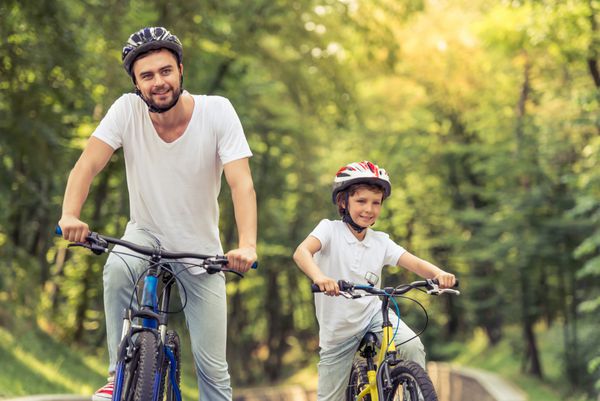 پدر جوان خوش تیپ و پسر کوچک نازش در حالی که در پارک دوچرخه سواری می کنند به جلو نگاه می کنند و لبخند می زنند