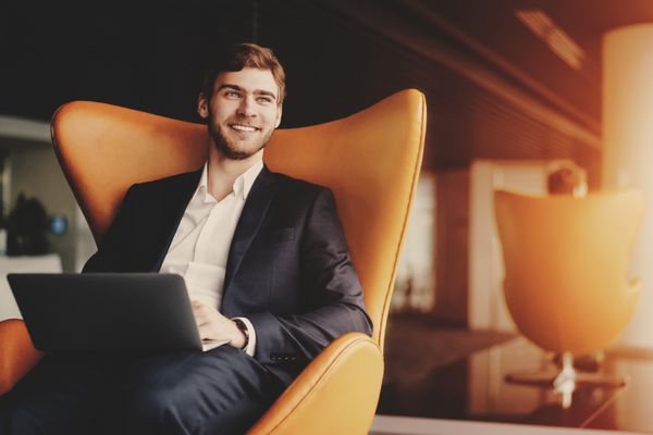 مرد موفق جوان خندان کارآفرین در مجموعه تجاری رسمی با ریش نشسته روی صندلی راحتی نارنجی با لپ تاپ در فضای داخلی اداری لوکس