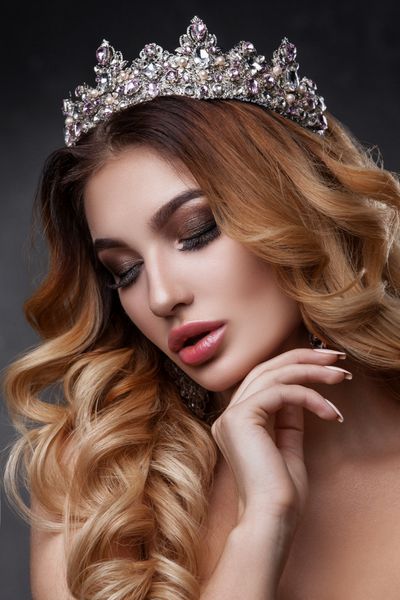 صورت زن زیبا با رنگ های آرایش زیبا تصویر ملکه موهای تیره تاجی روی سر پوست شفاف صورت زیبا لب های پرپشت پرتره در استودیو روی پس‌زمینه خاکستری گرفته شده است