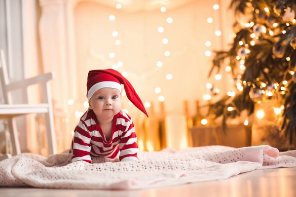 دختر بچه بامزه ای که کت و شلوار بابا نوئل به تن دارد و روی چراغ های کریسمس روی زمین می خزد نگاه کردن به دوربین فصل تعطیلی