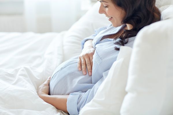 مفهوم بارداری استراحت مردم و انتظار - نمای نزدیک از زن باردار شاد و خندان که در رختخواب دراز کشیده و شکم خود را در خانه لمس می کند