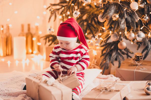 هدایای باز کریسمس دختر بچه ناز 1 ساله که روی زمین زیر درخت کریسمس در اتاق نشسته است فصل تعطیلی