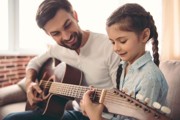 دختر کوچک ناز و پدر خوش تیپش در حالی که روی مبل خانه نشسته اند گیتار می زنند و لبخند می زنند
