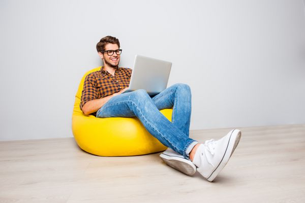 مرد جوان شادی که در پوف زرد نشسته و از لپ تاپ استفاده می کند