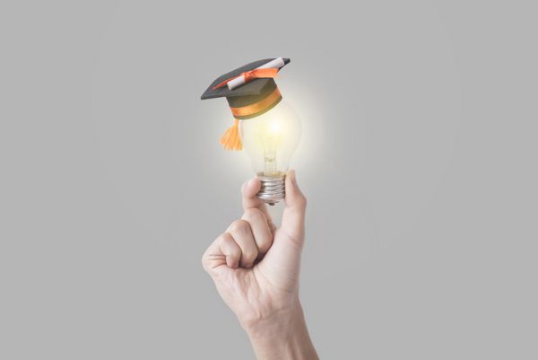 دست دانشجویی که لامپ در دست دارد با کلاه فارغ التحصیلی در پس زمینه خاکستری آموزش مفهومی ایده های جدید با نوآوری و خلاقیت