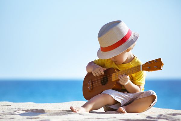 پسر کوچولو در پس زمینه ساحل دریا با گیتار یا یوکلله هاوایی می نوازد فضایی برای متن