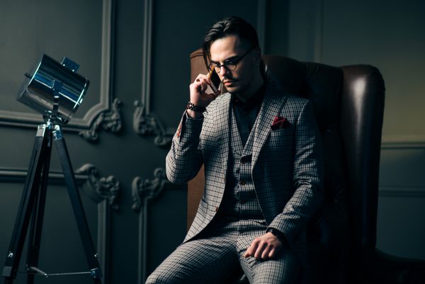 هیپستر جوان خوش تیپی که در استودیوی طراحی اتاق زیر شیروانی خود نشسته است از تلفن استفاده می کند و با کمک فن آوری های مدرن با مشتریان خود ارتباط برقرار می کند مرد شیک پوش با کت و شلوار مد روز