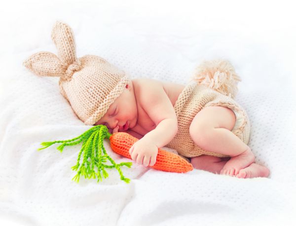 بسیار ناز نوزاد پسر دو هفته ای خندان با لباس بافتنی خرگوش کلاه با گوش خرگوش دم و اسباب بازی هویج خنده دار پرتره شیرین نوزاد تازه متولد شده در تخت خوابش