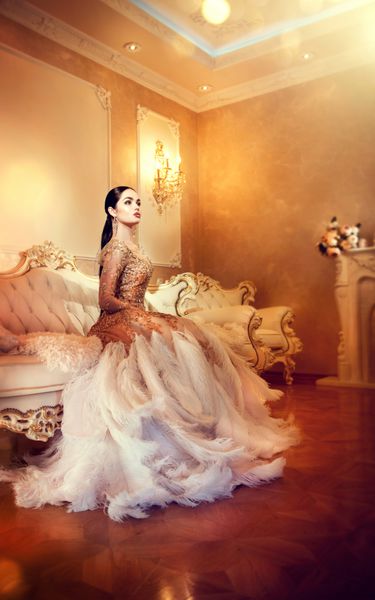 زن زیبا و زرق و برق دار با لباس شب زیبا که در اتاق داخلی به سبک لوکس نشسته و ژست گرفته است پرتره خانم شیک تمام قد با لباس طلایی