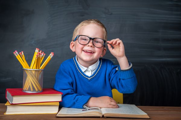 پسر باهوش و ناز با لیوانی با دست بالا پشت میز نشسته است کودک با تخته سیاه روی پس زمینه آماده پاسخگویی است آماده برای مدرسه بازگشت به مدرسه سیب و کتاب روی میز