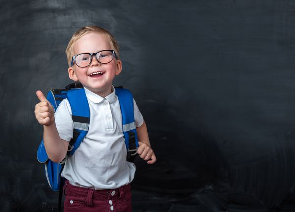 پسر خندان با عینک با شست بالا برای اولین بار به مدرسه می رود کودک با کیف و کتاب مدرسه کودک در داخل کلاس با پس زمینه تخته سیاه بازگشت به مدرسه