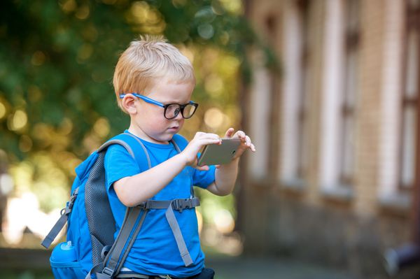 پسر باهوش زیبا با عینک با کیف مدرسه و گوشی هوشمند در دست در فضای باز آموزش در دبستان با تلفن همراه تکنولوژی جدید استفاده بیش از حد از تلفن همراه بازگشت به مدرسه