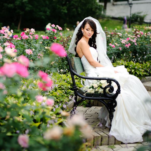 دختر زیبا در لباس عروسی که تنها در میان گل رز نشسته است