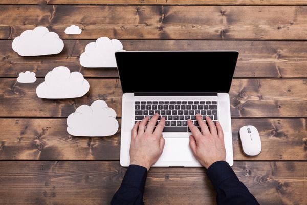 مفهوم فناوری محاسبات ابری با ابرهای کرکی سفید در کنار لپ تاپ دست‌های مرد در حال تایپ صفحه‌کلید در حال بارگذاری اطلاعات روی میز چوبی
