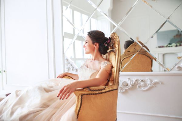 عروس با لباس زیبا روی مبل داخل خانه در فضای داخلی استودیو سفید مانند خانه نشسته است سبک عروسی مد روز عروسی