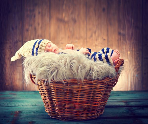 نوزاد دو هفته‌ای با لباس‌های خنده‌دار بافتنی خوابیده در سبد روی پس‌زمینه چوبی عکس استودیویی پرتره نوزاد شیرین روی پس‌زمینه روستایی چوبی