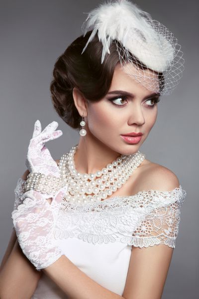 پرتره عروس عروسی از زن زیبای رترو خانمی با کلاه سفید و لباس‌های شیک مجموعه جواهرات مروارید مد روز را بر روی پس‌زمینه خاکستری نشان می‌دهد