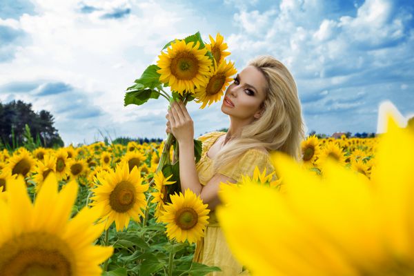 زن زیبا روی یک چمنزار با گل آفتابگردان ایستاده و با خوشحالی لبخند می زند