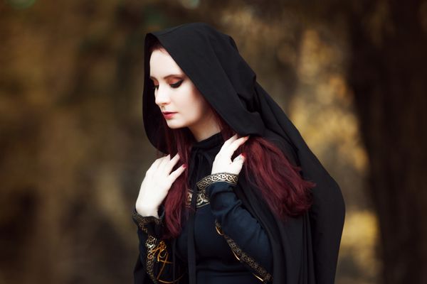زن جوان زیبا و مرموز در جنگل با شنل مشکی با کلاه تصویر جن جنگلی یا جادوگر