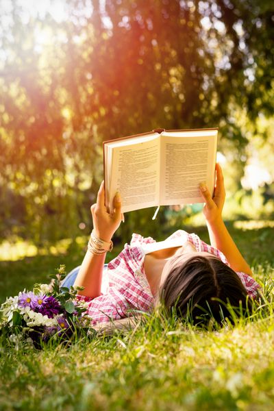 زن جوان در حال خواندن کتاب در پارک با گل