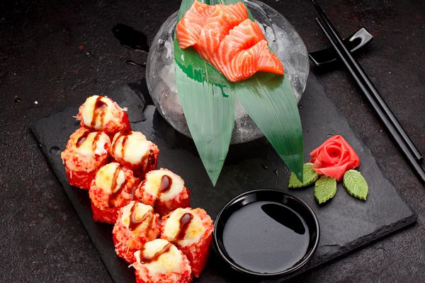 غذای ژاپنی رول سوشی پخته شده گرم و سیخ های ماهی قزل آلا روی صفحه سنگی روی زمینه بتنی سیاه