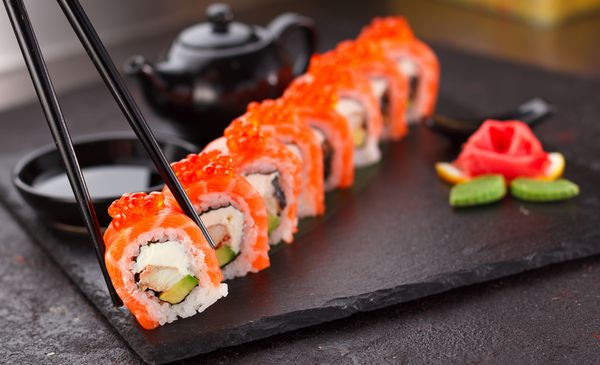 غذای ژاپنی سوشی ماهی قزل آلا در چاپستیک ها روی یک بشقاب سنگی روی زمینه بتنی می چرخد