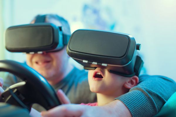 پدر و پسر با عینک واقعیت مجازی در حال بازی ویدیویی با چرخ های مسابقه ای در خانه