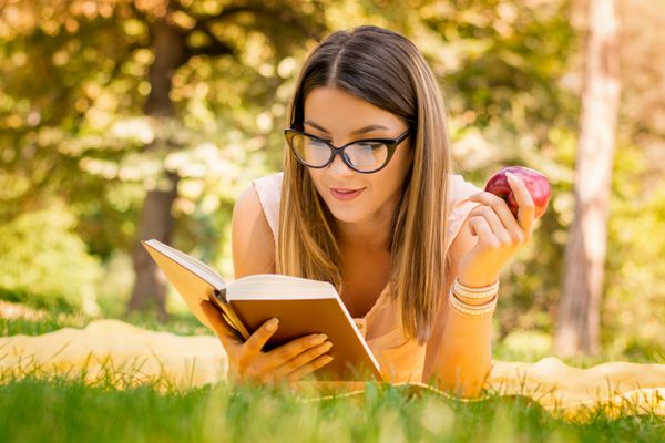 پرتره بیرونی زن جوان زیبا با کتاب خواندن سیب در پارک