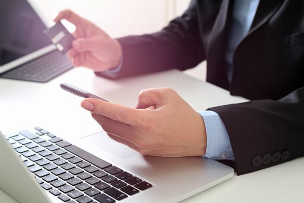 تاجر در حال خرید آنلاین کارت اعتباری با تلفن همراه و رایانه لپ تاپ روی میز مدرن