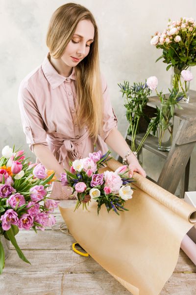 زنی در حال بسته بندی دسته گل صورتی بهار نمای بالا گل‌فروش جوان گل‌فروشی را در کارگاه روی زمینه چوبی مونتاژ می‌کند
