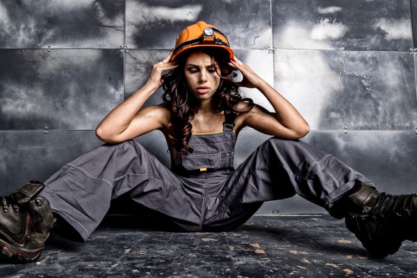 کارگر زیبا و خسته معدن با کلاه ایمنی نارنجی با چراغ قوه نشسته روی زمین در زمینه فولادی