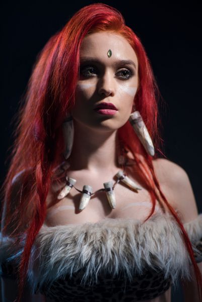 پرتره زیبا زن با موهای قرمز در عصر حجر لباس با carcanet دندان نما در دوربین در پس زمینه تاریک