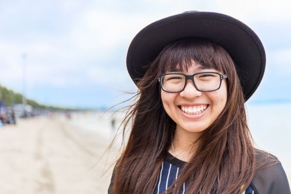 دختر نوجوان آسای با عینک و کلاه با پس زمینه ساحلی تار