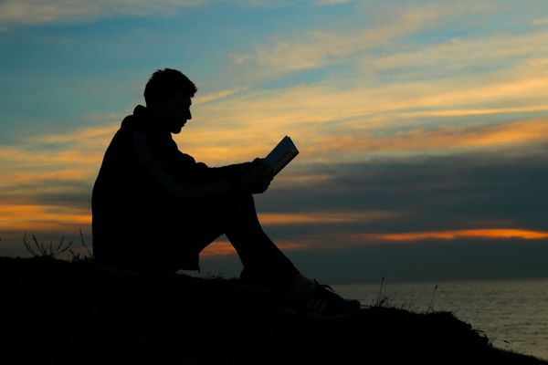 چهره ای از یک مرد نشسته بر روی موج شکن در شب در نزدیکی دریا کتاب می خواند