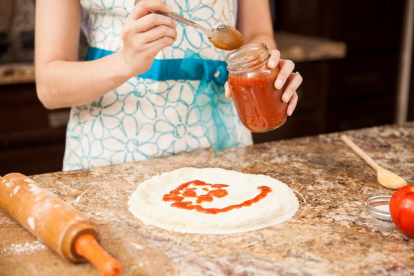 بستن یک زن با یک پریز با افزودن سس گوجه فرنگی به پیتزا خانگی خود در آشپزخانه