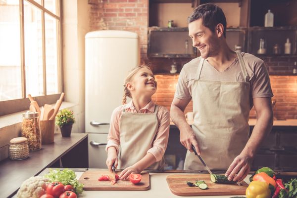 دختر کوچک ناز و پدرخوانده او سبزیجات را بریده و لبخند می زنند در حالی که آشپزخانه را در خانه پخت و پز می کنند