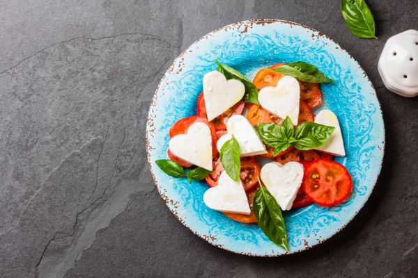 سالاد ایتالیایی مدیترانه ای Caprese با گوجه فرنگی و پنیر مزارعلا به عنوان قلب و ریحان بر روی صفحه آبی بر روی زمینه تخته سنگ برش داده شده منوی روزانه نمای بالا