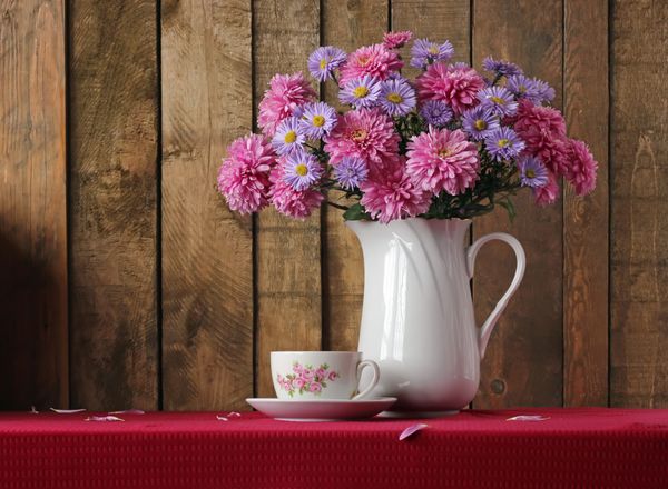 زندگی با یک دسته گل های بنفش و صورتی پاییز و یک فنجان یکپارچه بر روی میز با یک پارچه قرمز در برابر تخته ها کارت تبریک