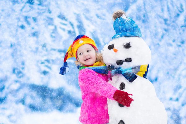 دختر خنده دار کمی کودک نو پا در یک کلاه رنگارنگ و کت گرم با مرد برفی کودکان در زمستان در خارج از منزل بازی می کنند بچه ها سرگرم کننده در زمان کریسمس کودک آدم برفی در Xmas