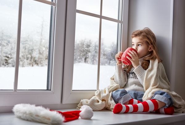 دختر کوچک نشسته توسط پنجره با یک فنجان نوشیدنی و به دنبال جنگل زمستان