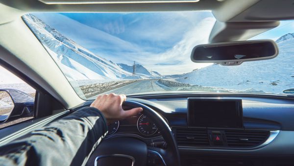 نمایش از ماشین لوکس در داخل و بخشی از صفحه نمایش داخلی جیپیاس با دست راننده مرد در فرمان در طول روز آفتابی برفی در جاده مستقیم یخی با کوه های برفی در پس زمینه